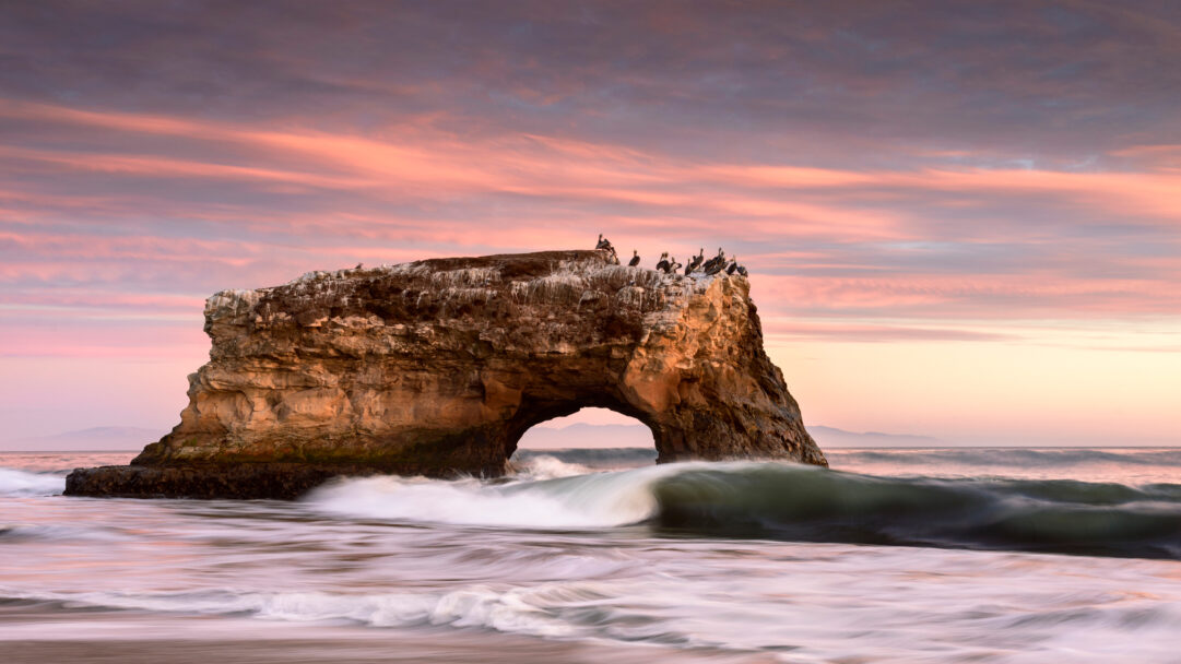 A breathtaking 4K wallpaper showcasing Natural Bridges State Beach in California at sunset, with majestic ocean waves crashing around the iconic sea arch. The sky glows with serene pink and purple hues, reflecting a tranquil and beautiful natural scene.