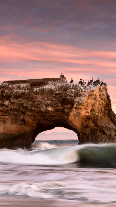 A breathtaking 4K wallpaper showcasing Natural Bridges State Beach in California at sunset, with majestic ocean waves crashing around the iconic sea arch. The sky glows with serene pink and purple hues, reflecting a tranquil and beautiful natural scene.