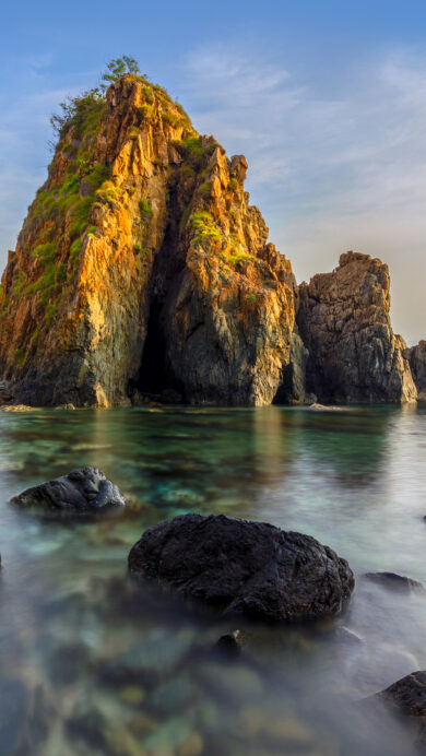 A breathtaking 4K wallpaper presents the majestic Nha Phu Bay in Nha Trang, Vietnam, showcasing a dramatic rocky seascape where towering formations meet calm, clear waters. The warm, golden light of dawn bathes the rugged peaks in a vibrant glow, sharply contrasting with the silky smooth, mist-like effect of the gently moving sea over dark, ancient rocks.