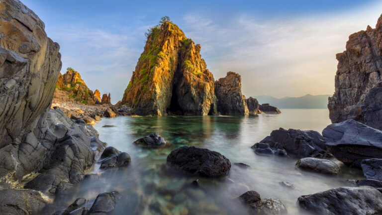 A breathtaking 4K wallpaper presents the majestic Nha Phu Bay in Nha Trang, Vietnam, showcasing a dramatic rocky seascape where towering formations meet calm, clear waters. The warm, golden light of dawn bathes the rugged peaks in a vibrant glow, sharply contrasting with the silky smooth, mist-like effect of the gently moving sea over dark, ancient rocks.