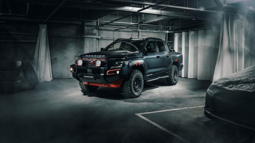 An imposing 4K wallpaper presenting a matte black Nissan Navara Warrior pickup truck, parked dramatically under moody lighting in an industrial-style studio. Its powerful stance is accentuated by bright orange accents and prominent LED lights, cutting through a subtle atmospheric haze to convey a sense of raw capability and style.