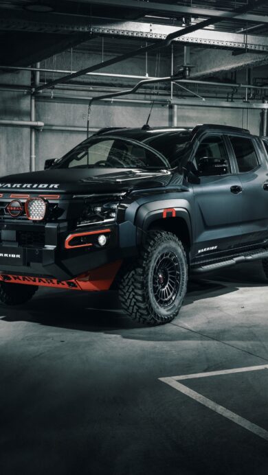 An imposing 4K wallpaper presenting a matte black Nissan Navara Warrior pickup truck, parked dramatically under moody lighting in an industrial-style studio. Its powerful stance is accentuated by bright orange accents and prominent LED lights, cutting through a subtle atmospheric haze to convey a sense of raw capability and style.