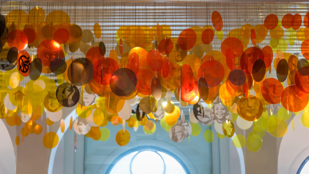 A vibrant 4K wallpaper showcasing the Nobel Peace Center's art installation in Oslo, where numerous translucent orange and yellow discs hang suspended from the ceiling. Light filters through the overlapping discs, revealing subtle etched portraits of Nobel laureates and symbols of peace, creating an inspiring glow against the architectural backdrop.