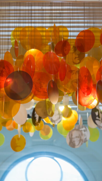 A vibrant 4K wallpaper showcasing the Nobel Peace Center's art installation in Oslo, where numerous translucent orange and yellow discs hang suspended from the ceiling. Light filters through the overlapping discs, revealing subtle etched portraits of Nobel laureates and symbols of peace, creating an inspiring glow against the architectural backdrop.