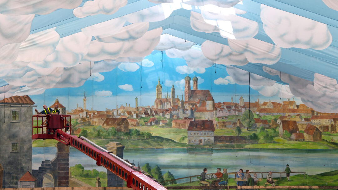 A grand 4K wallpaper depicting the elaborate interior of an Oktoberfest beer tent, featuring a vast painted mural of a historic Munich cityscape under an artificial sky ceiling. Billowing fabric clouds drift across the blue sky ceiling above the detailed townscape and a serene river, while two workers on a bright red boom lift add a dynamic human element to the immersive, festive scene.