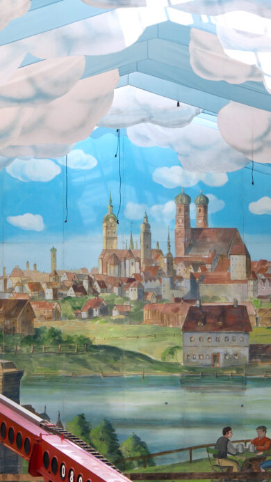 A grand 4K wallpaper depicting the elaborate interior of an Oktoberfest beer tent, featuring a vast painted mural of a historic Munich cityscape under an artificial sky ceiling. Billowing fabric clouds drift across the blue sky ceiling above the detailed townscape and a serene river, while two workers on a bright red boom lift add a dynamic human element to the immersive, festive scene.
