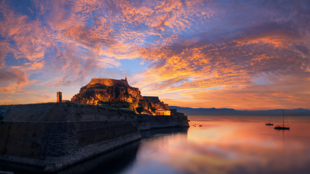 A breathtaking 4K wallpaper features the Old Fortress of Corfu in Greece standing majestically by the sea, silhouetted against a dramatic sunset sky. Fiery orange and deep purple clouds fill the vast sky, reflecting intensely on the calm water where two sailboats float, creating a truly spectacular and serene scene.
