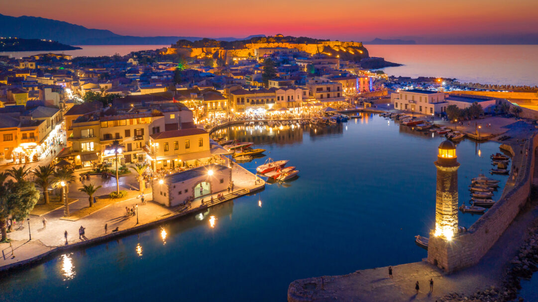 A mesmerizing 4K wallpaper captures the Old Venetian Harbor of Rethymno, Crete, stretching along the coastline as a vibrant cityscape at sunset. The iconic lighthouse beams brightly, its warm glow along with numerous building lights reflecting vividly on the deep blue harbor waters beneath a breathtaking orange and purple twilight sky.