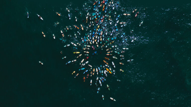 A captivating 4K wallpaper showcasing a large 'Paddle Out For Unity' event in the dark teal ocean off San Diego, California, where hundreds of surfers and paddleboarders are closely gathered. The aerial view emphasizes the dense, colorful cluster of boards forming a striking visual of solidarity against the deep, shimmering expanse of the Pacific Ocean.