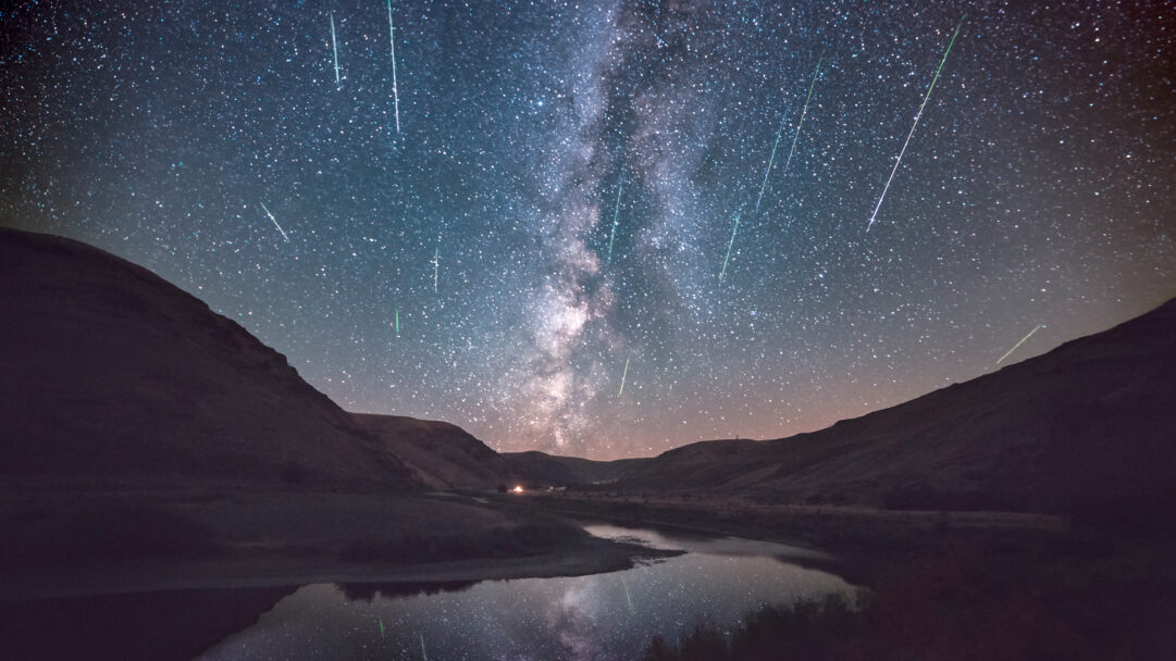 A celestial 4K wallpaper depicting the Perseid Meteor Shower and the majestic Milky Way arching over Cottonwood Canyon State Park in Oregon. Numerous bright meteors streak across the dark night sky, their glowing trails and the Milky Way's band reflecting in the winding river below, creating a profound sense of cosmic wonder.