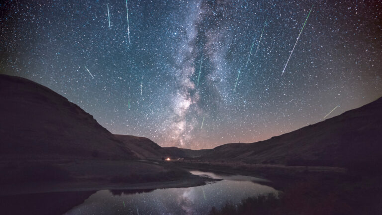 A celestial 4K wallpaper depicting the Perseid Meteor Shower and the majestic Milky Way arching over Cottonwood Canyon State Park in Oregon. Numerous bright meteors streak across the dark night sky, their glowing trails and the Milky Way's band reflecting in the winding river below, creating a profound sense of cosmic wonder.