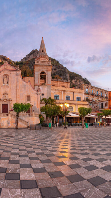 An enchanting 4K wallpaper showcasing the historic Piazza IX Aprile in Taormina, Sicily, Italy, at twilight. Illuminated historic buildings and the square's distinctive checkered pavement glow under a dramatic twilight sky painted with soft pink and blue hues.