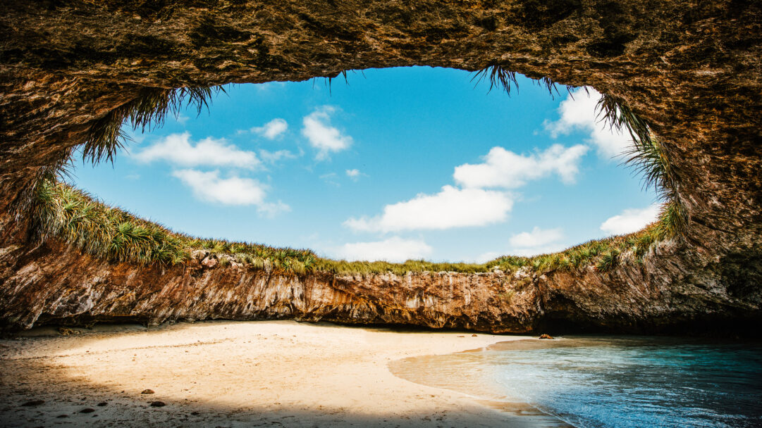 A captivating 4K wallpaper of Playa del Amor Hidden Beach in Mexico's Marietas Islands, nestled within a dramatic open grotto formed by rock and foliage. The brilliant sun shines through the circular opening, illuminating the golden sand and clear turquoise waters for a tranquil escape.