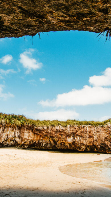 A captivating 4K wallpaper of Playa del Amor Hidden Beach in Mexico's Marietas Islands, nestled within a dramatic open grotto formed by rock and foliage. The brilliant sun shines through the circular opening, illuminating the golden sand and clear turquoise waters for a tranquil escape.