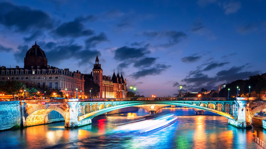 An enchanting 4K wallpaper captures the Pont d'Arcole Bridge spanning the Seine River in Paris at night, flanked by beautifully lit historic buildings. Vibrant city lights cast colorful reflections on the rippling water, with the blurred trail of a boat adding dynamic energy to the nocturnal scene.
