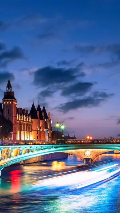 An enchanting 4K wallpaper captures the Pont d'Arcole Bridge spanning the Seine River in Paris at night, flanked by beautifully lit historic buildings. Vibrant city lights cast colorful reflections on the rippling water, with the blurred trail of a boat adding dynamic energy to the nocturnal scene.