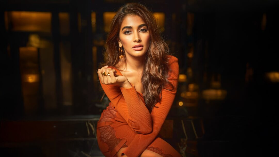A captivating 4K wallpaper featuring Pooja Hegde, dressed in a form-fitting orange knit dress, posing confidently in a dimly lit, luxurious indoor setting. The striking contrast between her vibrant orange attire and the dark, blurred background highlights her intense gaze and the subtle gold accents of her jewelry, creating a mood of dramatic allure.