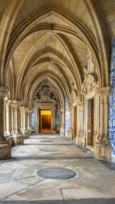 An exquisite 4K wallpaper captures the long, vaulted cloister of the Porto Cathedral in Portugal, featuring rows of ancient stone arches and columns receding into the distance. One entire wall is adorned with striking blue and white azulejo tiles, depicting intricate historical scenes that are beautifully illuminated by soft light filtering through the opposite arcade.