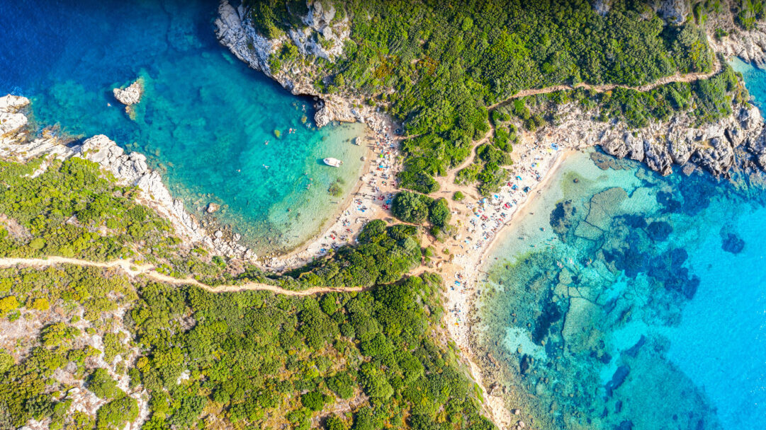 A captivating 4K wallpaper offers an aerial view of Porto Timoni Beach in Corfu, Greece, where two crescent beaches meet to form a striking natural double cove. The brilliantly clear turquoise waters, dotted with swimmers and small boats, are framed by lush green hillsides, creating an inviting and serene tropical escape.