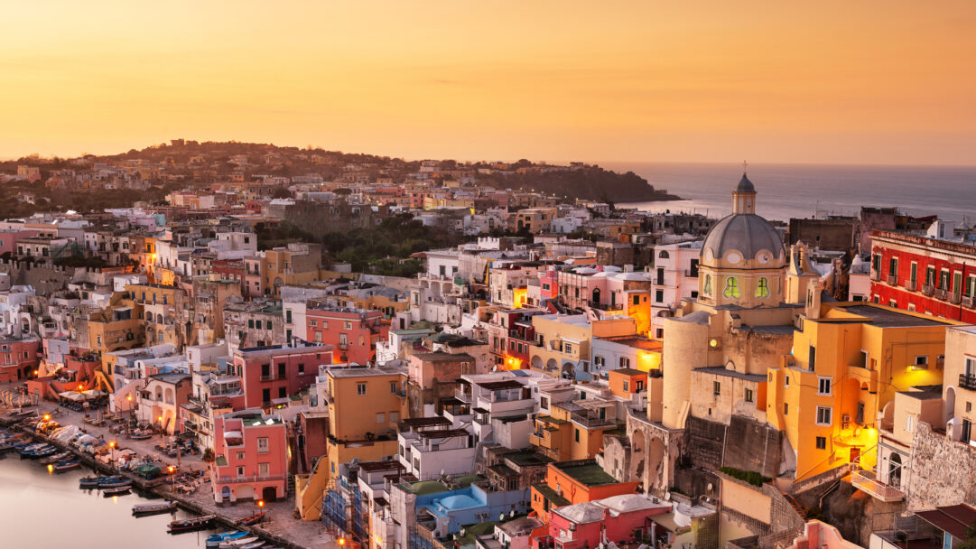 A captivating 4K wallpaper showcasing Procida, Italy, a vibrant coastal town nestled along the shimmering sea as the sun sets. The golden light bathes the densely packed, pastel-colored buildings, illuminating a prominent church dome and creating a warm, idyllic evening mood.