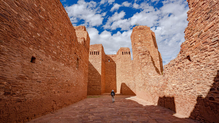 A historic 4K wallpaper capturing the towering red stone ruins of the Quarai Mission Church at Salinas Pueblo Missions National Monument, with a lone person walking on a textured stone path. Dramatic sunlight casts deep shadows across the ancient structure, accentuating its rich red hues against a brilliant blue sky dotted with fluffy white clouds and conveying a sense of enduring grandeur.