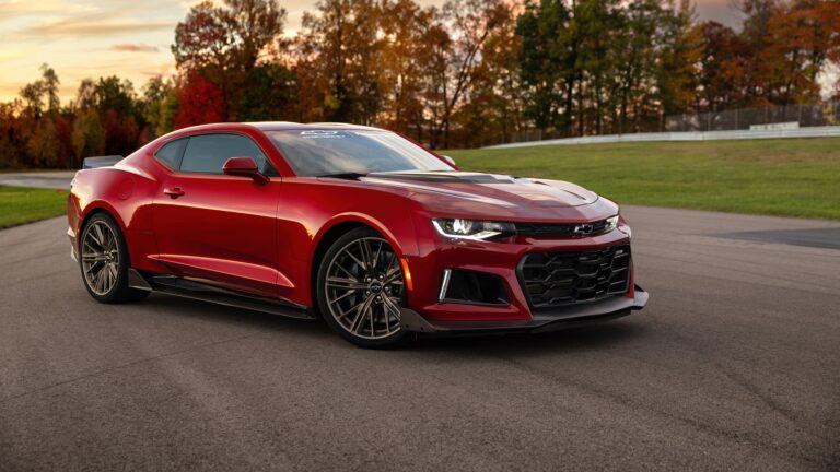 A spectacular 4K wallpaper showcasing a vibrant red Chevrolet Camaro ZL1 parked on a racetrack. Its sleek lines gleam under the warm autumn sunset, casting a golden glow on the car's rich red paint and highlighting the fiery foliage in the background.