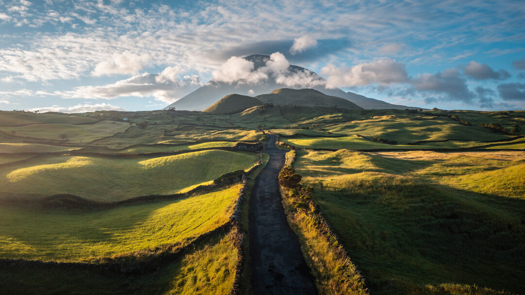 A magnificent 4K wallpaper features a winding road through the vibrant green hills of Portugal, stretching towards the imposing Mount Pico. Golden light illuminates the rolling landscape, highlighting the intricate stone walls and dramatic cloud formations around the distant peak.