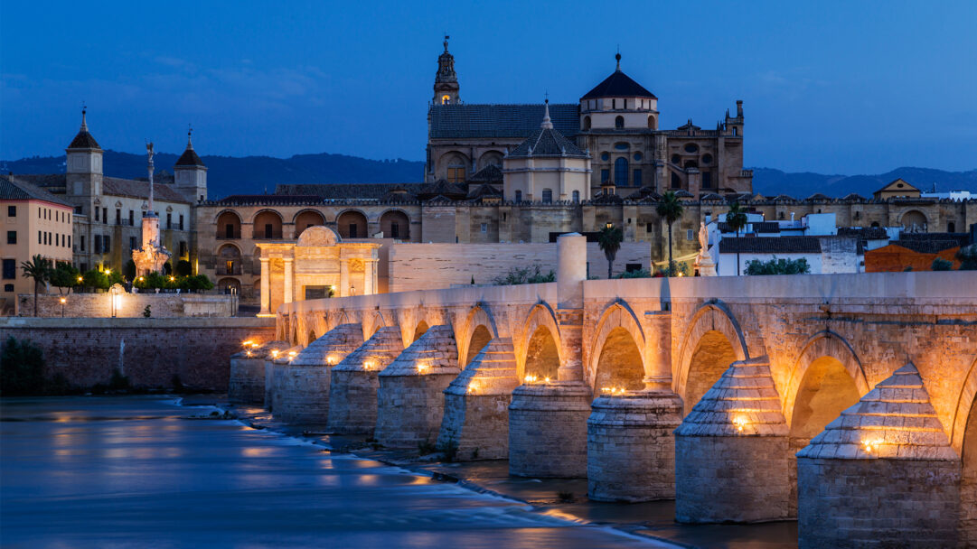 An enchanting 4K wallpaper captures the illuminated Roman Bridge in Cordoba, Spain, stretching across a river at dusk with the historic Mezquita-Cathedral dominating the city skyline. The bridge's golden arches glow warmly against the tranquil, deep blue water and twilight sky, creating a serene and timeless atmosphere.