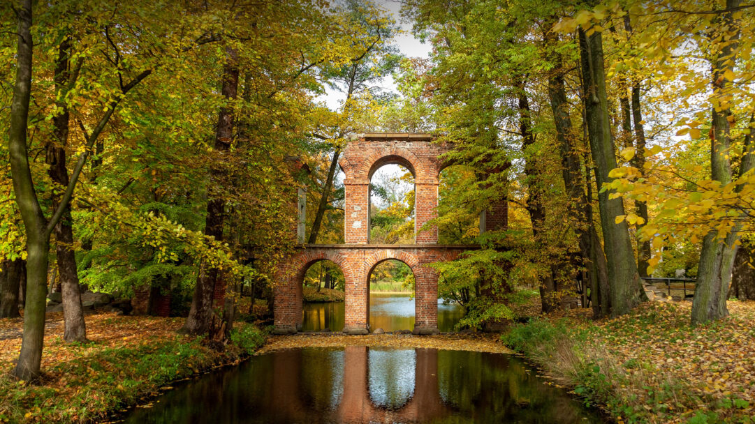 An evocative 4K wallpaper capturing a Roman-inspired brick aqueduct nestled within Arkadia Park, Poland, during the vibrant autumn season. Its weathered brick arches are beautifully framed by a canopy of yellow and green autumn leaves, with the structure and foliage reflecting perfectly in the still, dark water below.
