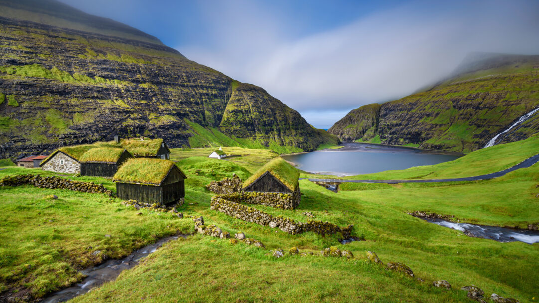 An immersive 4K wallpaper displays traditional turf roof houses nestled in the dramatic, verdant fjord landscape of Saksun, Faroe Islands, Denmark. Lush green turf roofs blend seamlessly with the steep, moss-covered mountainsides under a sky where mist delicately cloaks the peaks, reflecting a profound sense of tranquil isolation.