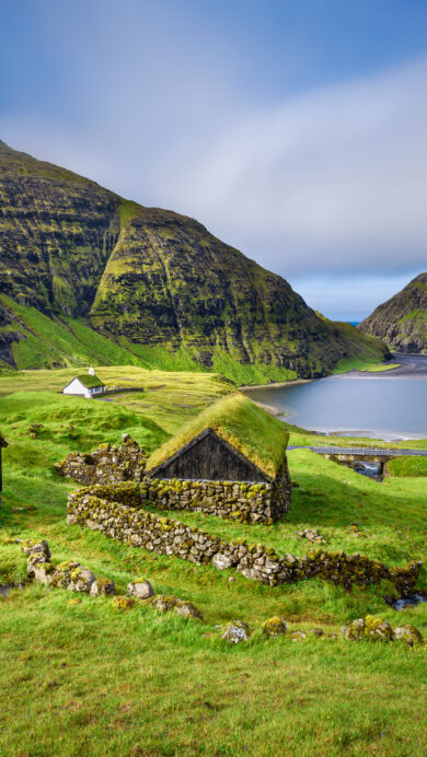 An immersive 4K wallpaper displays traditional turf roof houses nestled in the dramatic, verdant fjord landscape of Saksun, Faroe Islands, Denmark. Lush green turf roofs blend seamlessly with the steep, moss-covered mountainsides under a sky where mist delicately cloaks the peaks, reflecting a profound sense of tranquil isolation.