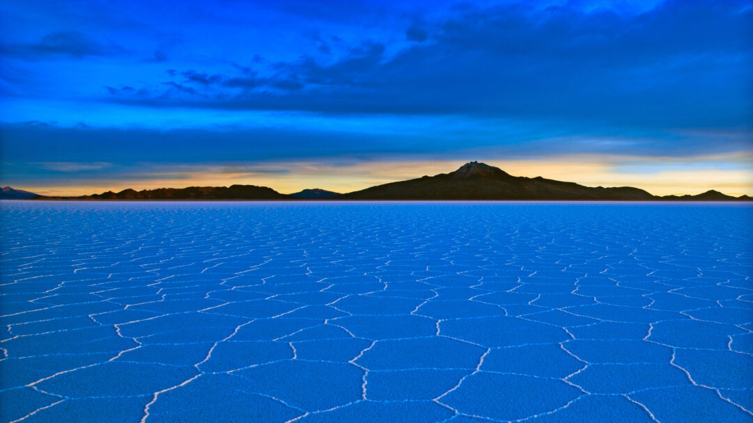 An immersive 4K wallpaper showcasing the Salar de Uyuni in Bolivia, featuring a vast, intensely blue salt flat stretching towards distant mountains under a dramatic sky. The captivating hexagonal patterns on the vivid azure ground reflect the deep blue overhead, creating a serene and otherworldly landscape touched by a subtle golden glow on the horizon.