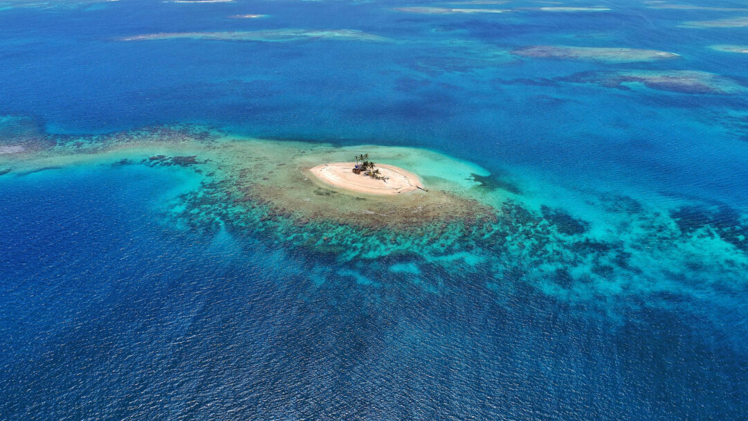 A captivating 4K wallpaper reveals a tiny, pristine tropical island in the San Blas Islands, Panama, isolated amidst a vast expanse of clear blue ocean. The scene is defined by the mesmerizing transition from the island’s sandy shores and scattered palm trees to the crystalline turquoise shallows, deepening into profound oceanic blues that reveal underwater reefs.