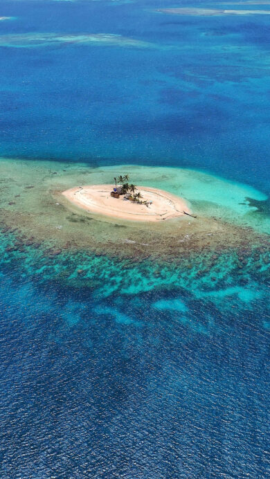A captivating 4K wallpaper reveals a tiny, pristine tropical island in the San Blas Islands, Panama, isolated amidst a vast expanse of clear blue ocean. The scene is defined by the mesmerizing transition from the island’s sandy shores and scattered palm trees to the crystalline turquoise shallows, deepening into profound oceanic blues that reveal underwater reefs.