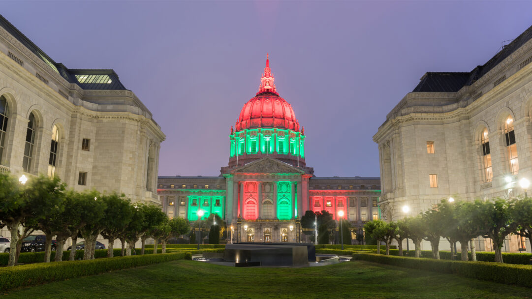 A commemorative 4K wallpaper beautifully displays San Francisco City Hall, a majestic Beaux-Arts building, illuminated in vibrant red and green lights for Juneteenth against a soft twilight sky. The striking color scheme vividly adorns the iconic dome and facade, casting a celebratory glow over the tranquil surrounding lawns and creating a powerful visual tribute.