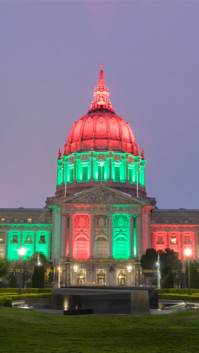 A commemorative 4K wallpaper beautifully displays San Francisco City Hall, a majestic Beaux-Arts building, illuminated in vibrant red and green lights for Juneteenth against a soft twilight sky. The striking color scheme vividly adorns the iconic dome and facade, casting a celebratory glow over the tranquil surrounding lawns and creating a powerful visual tribute.