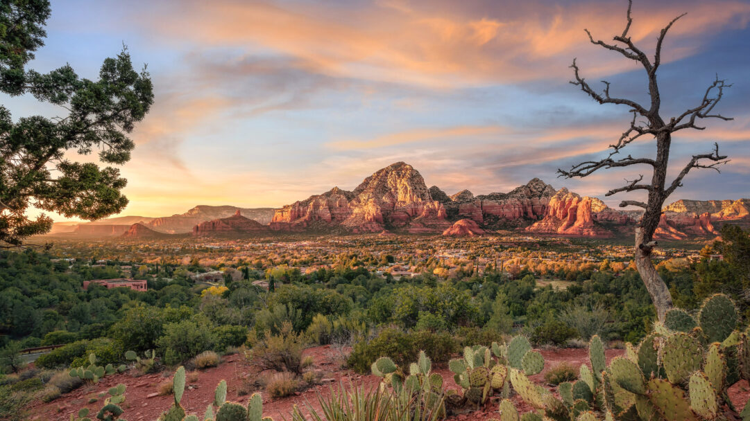 An exquisite 4K wallpaper capturing the majestic red rock formations of Sedona, Arizona, dominating a sprawling desert landscape with a vibrant town nestled beneath. The warm golden light of the sunset dramatically illuminates the textured red rocks and the lush green desert landscape, under a sky painted with soft orange and blue hues.