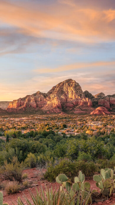 An exquisite 4K wallpaper capturing the majestic red rock formations of Sedona, Arizona, dominating a sprawling desert landscape with a vibrant town nestled beneath. The warm golden light of the sunset dramatically illuminates the textured red rocks and the lush green desert landscape, under a sky painted with soft orange and blue hues.
