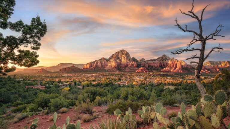 An exquisite 4K wallpaper capturing the majestic red rock formations of Sedona, Arizona, dominating a sprawling desert landscape with a vibrant town nestled beneath. The warm golden light of the sunset dramatically illuminates the textured red rocks and the lush green desert landscape, under a sky painted with soft orange and blue hues.