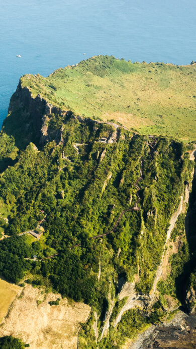 An awe-inspiring 4K wallpaper of Seongsan Ilchulbong Tuff Cone, an emerald-green volcanic crater island rising majestically from the vibrant blue waters off Jeju Island, South Korea. Sunlight brilliantly illuminates the cone's verdant crater rim and its dramatic, textured cliffs plunging into the clear azure sea, highlighting a serene expanse where a small boat leaves a delicate wake.