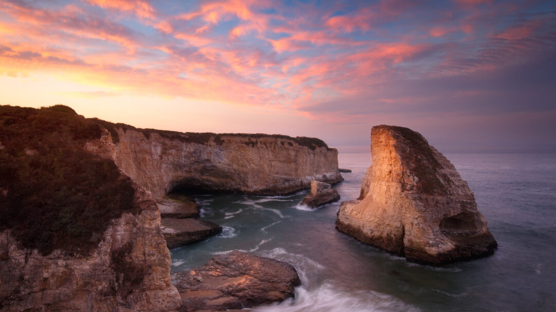 A captivating 4K wallpaper showcasing Shark Fin Cove and its majestic ocean cliffs during sunset in Davenport, California. The sky above explodes with magnificent pink and orange clouds, casting a warm, ethereal glow over the distinctive shark fin rock formation and the gentle, long-exposure waves below.