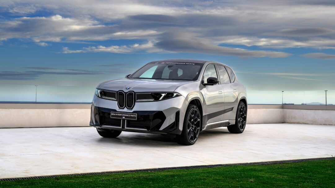 A dynamic 4K wallpaper presenting a silver BMW iX M Performance SUV parked on a light-colored paved terrace overlooking a calm blue ocean, under a dramatic sky filled with layered clouds. Its futuristic design and metallic finish, enhanced by M Performance parts, reflect the soft light, emphasizing its sleek power and sophisticated presence against the natural coastal backdrop.