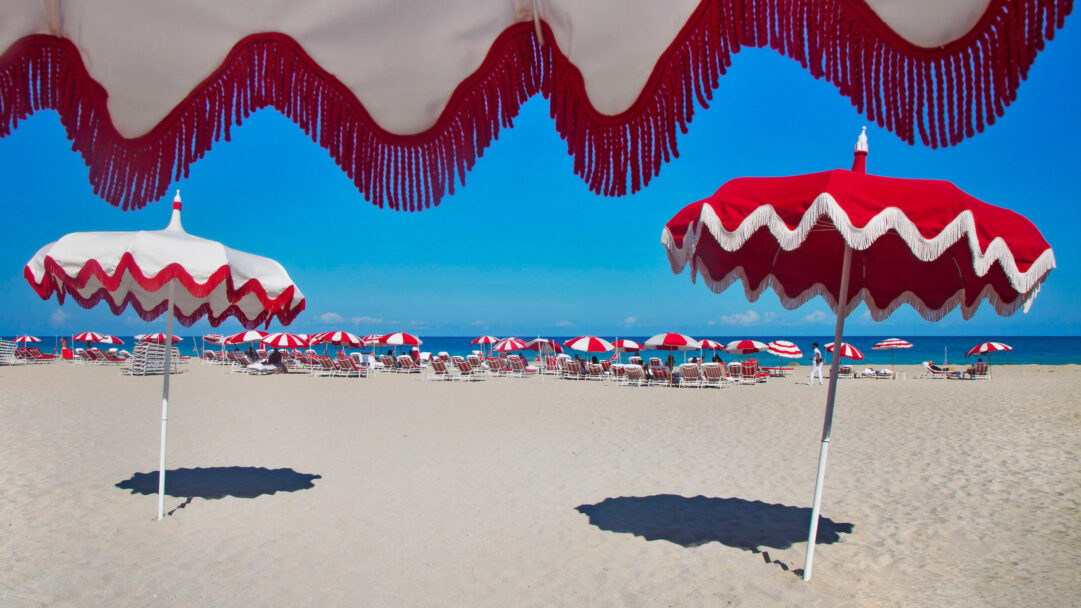 An iconic 4K wallpaper presents South Beach, Miami Beach, Florida, bustling with red and white umbrellas and lounge chairs lining the vast sandy beach stretching to the clear blue ocean. The striking red and white patterns of the umbrellas create a cheerful rhythm across the sun-drenched sand, vividly contrasted by the brilliant blue sky and calm ocean waters.