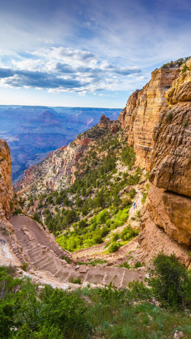 A majestic 4K wallpaper captures the South Kaibab Trail as it gracefully descends through the colossal geological formations of Grand Canyon National Park in Arizona. The deep reds and oranges of the canyon walls are vividly contrasted by patches of green foliage and the hazy blue depths of the distant canyon under a dramatic, cloud-streaked sky.