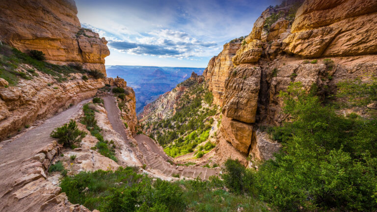 A majestic 4K wallpaper captures the South Kaibab Trail as it gracefully descends through the colossal geological formations of Grand Canyon National Park in Arizona. The deep reds and oranges of the canyon walls are vividly contrasted by patches of green foliage and the hazy blue depths of the distant canyon under a dramatic, cloud-streaked sky.