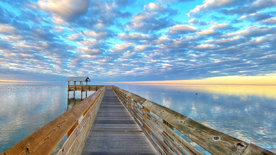 A mesmerizing 4K wallpaper showcasing a long wooden pier extending into the calm waters of South Padre Island, Texas. The sunset sky, filled with layered blue and soft golden clouds, casts a serene glow that reflects beautifully across the tranquil water, enhancing the scene's peaceful mood.