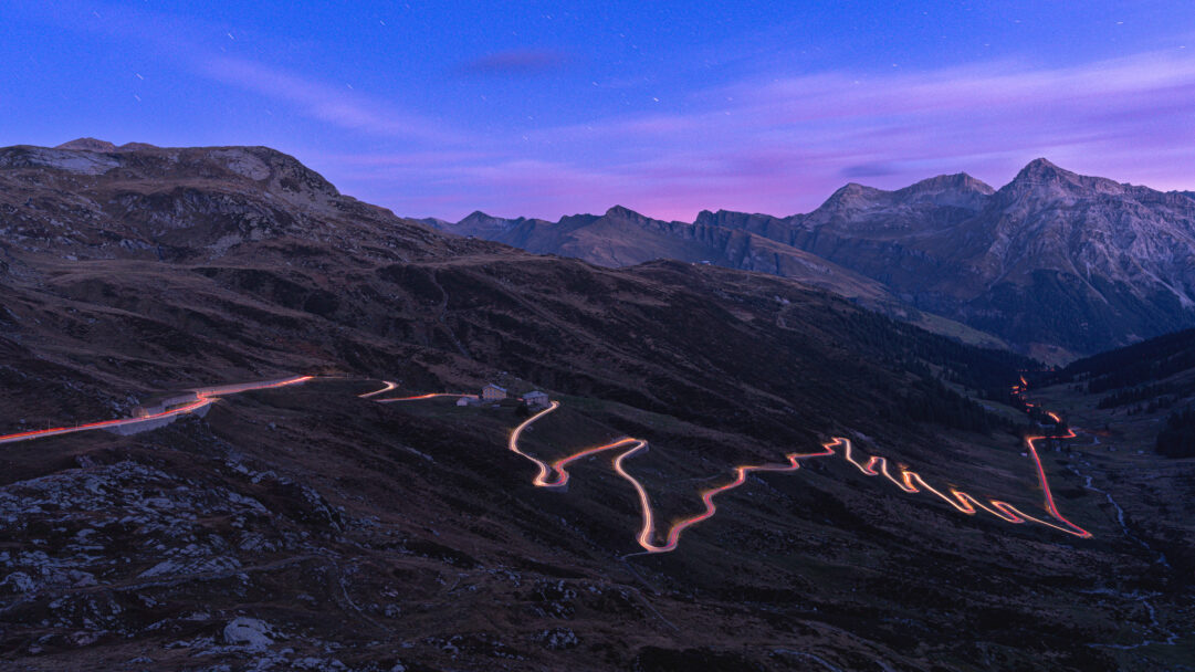 A captivating 4K wallpaper showcasing the winding mountain road of Splügen Pass in Switzerland at night. Vibrant red and yellow light trails from vehicles dramatically carve through the dark, rugged landscape, illuminated by the deep blue and purple twilight sky scattered with faint stars.