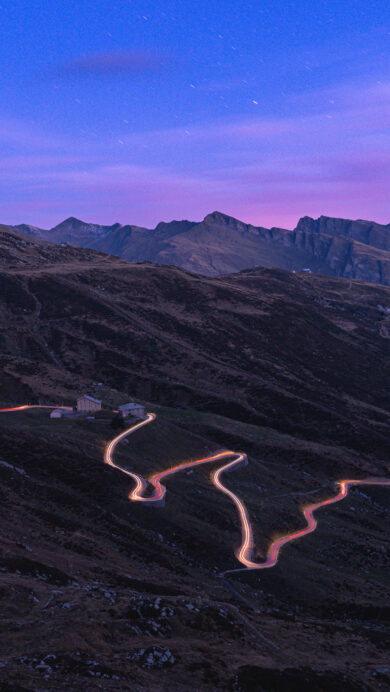 A captivating 4K wallpaper showcasing the winding mountain road of Splügen Pass in Switzerland at night. Vibrant red and yellow light trails from vehicles dramatically carve through the dark, rugged landscape, illuminated by the deep blue and purple twilight sky scattered with faint stars.