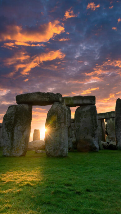 A breathtaking 4K wallpaper featuring Stonehenge at sunrise on Salisbury Plain, Wiltshire, England. The rising sun dramatically bursts between the ancient monolithic stones, painting the vast sky with vivid orange and deep blue clouds while illuminating the monument and foreground with a golden glow.