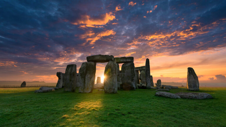 A breathtaking 4K wallpaper featuring Stonehenge at sunrise on Salisbury Plain, Wiltshire, England. The rising sun dramatically bursts between the ancient monolithic stones, painting the vast sky with vivid orange and deep blue clouds while illuminating the monument and foreground with a golden glow.